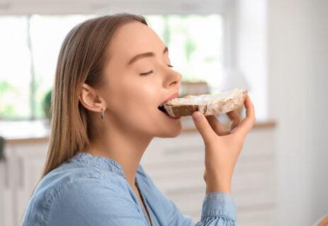 Beautiful young happy woman eating tasty toast with fresh butter in kitchen at home